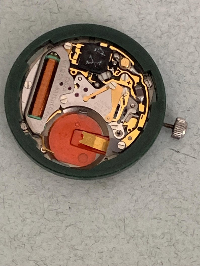 May include: A close-up view of a Citizen watch movement. The watch mechanism is housed in a dark green circular case. The internal components include a copper-colored coil, a red battery, and gold-colored gears and levers. The watch's crown is visible on the right side.
