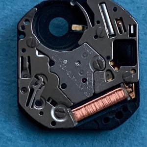 May include: Close-up of a watch movement with intricate mechanical components. The movement features silver-colored metal parts, a black circular component, and a copper-colored coil. The text "JAPAN" and "C 7511" are visible.