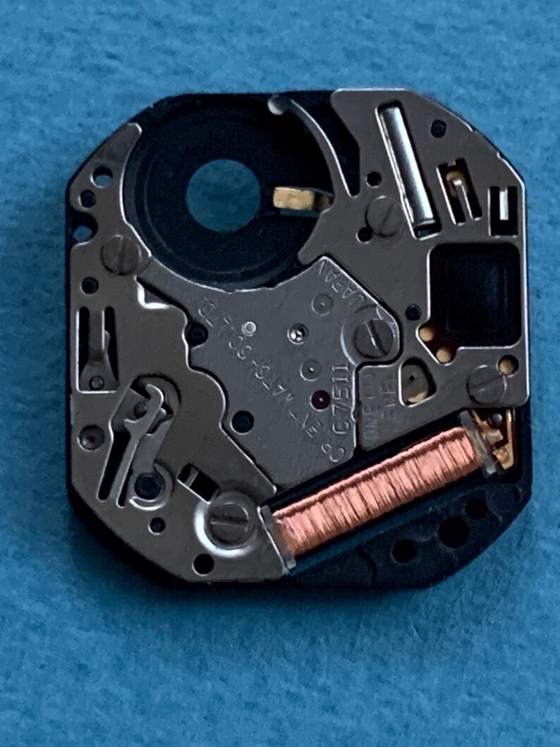 May include: Close-up of a watch movement with intricate mechanical components. The movement features silver-colored metal parts, a black circular component, and a copper-colored coil. The text "JAPAN" and "C 7511" are visible.