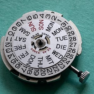 May include: A watch movement with a white dial displaying the days of the week and dates. The dial has black numerals and text, with a silver-colored metal frame and a small crown. The text includes "SUN", "MON", "TUE", "WED", "THU", "FRI", and "SAT".