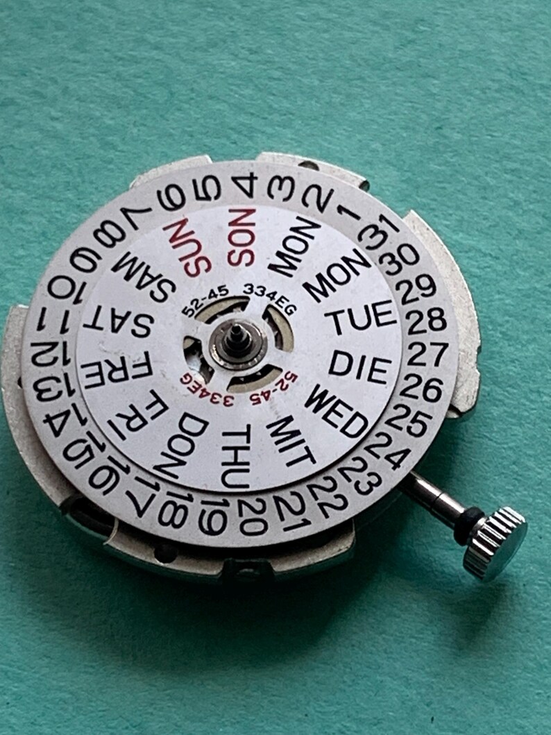 May include: A watch movement with a white dial displaying the days of the week and dates. The dial has black numerals and text, with a silver-colored metal frame and a small crown. The text includes "SUN", "MON", "TUE", "WED", "THU", "FRI", and "SAT".