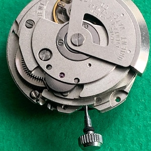May include: Close-up of a mechanical watch movement with intricate gears and components. The silver-toned parts are arranged in a circular pattern, with the text "ORIENT WATCH" visible. A small, silver crown is attached to the movement.