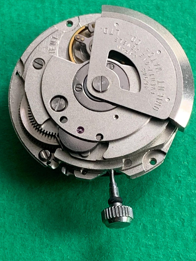 May include: Close-up of a mechanical watch movement with intricate gears and components. The silver-toned parts are arranged in a circular pattern, with the text "ORIENT WATCH" visible. A small, silver crown is attached to the movement.