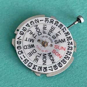 May include: A white watch calendar dial with black numerals and day abbreviations. The day of the week "SUN" is in red. A small screw extends from the dial. The dial has the text "58-45 334EG".