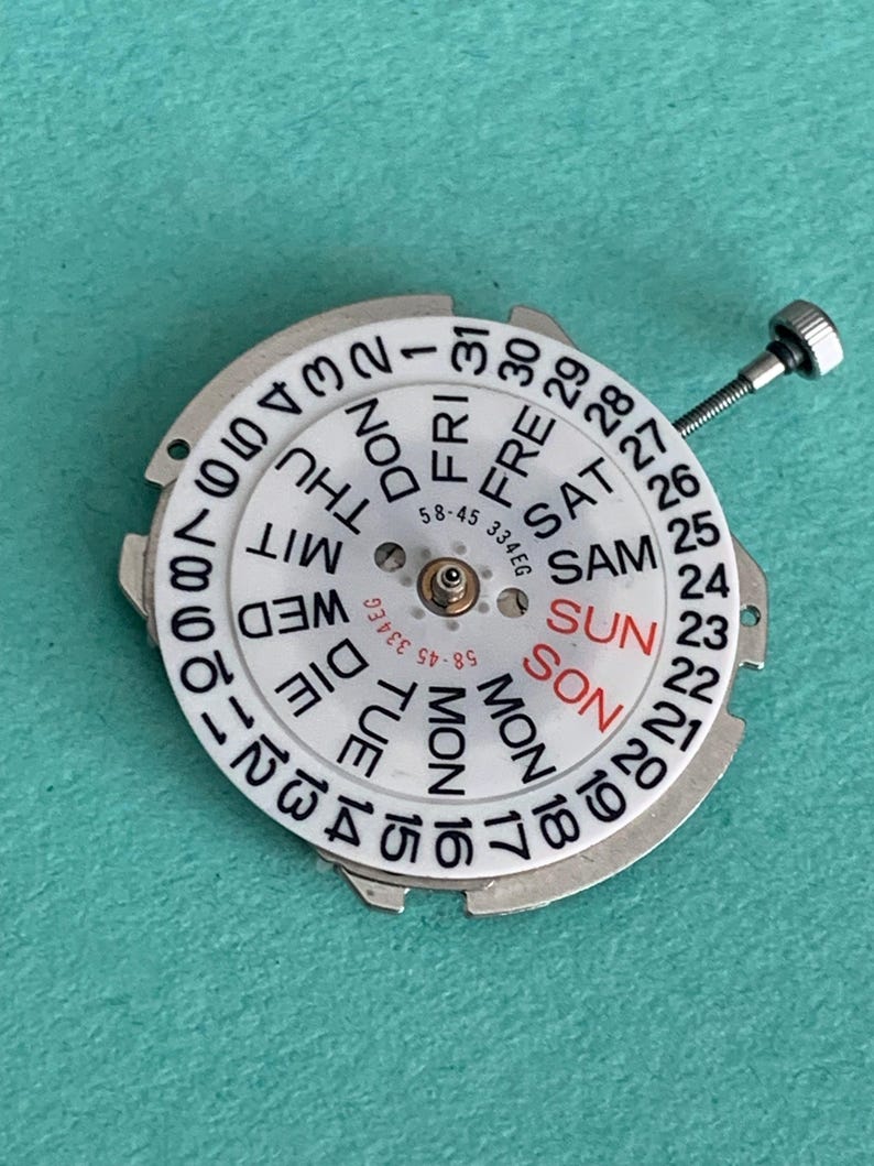 May include: A white watch calendar dial with black numerals and day abbreviations. The day of the week "SUN" is in red. A small screw extends from the dial. The dial has the text "58-45 334EG".