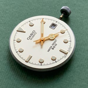 K&ouml;nnte beinhalten: Ein Casio Quartz Zifferblatt mit goldfarbenen Zeigern und Markierungen, einem Datumsfenster und dem Text "WATER RESIST 50M". Das Zifferblatt ist wei&szlig; mit einer goldfarbenen L&uuml;nette und einer silbernen Krone.