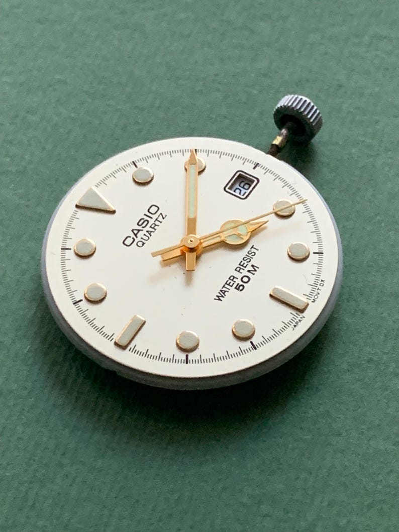 K&ouml;nnte beinhalten: Ein Casio Quartz Zifferblatt mit goldfarbenen Zeigern und Markierungen, einem Datumsfenster und dem Text "WATER RESIST 50M". Das Zifferblatt ist wei&szlig; mit einer goldfarbenen L&uuml;nette und einer silbernen Krone.