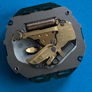 May include: A detailed view of a watch movement, featuring gold-toned gears, a copper coil, and silver and green metal parts. The watch mechanism has the text "N. JEWEL WATCH CO. JAPAN" and "2110A".