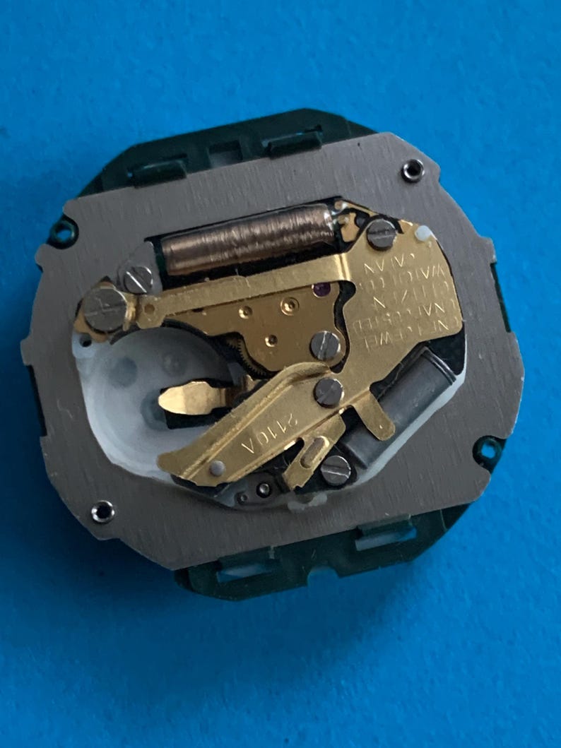 May include: A detailed view of a watch movement, featuring gold-toned gears, a copper coil, and silver and green metal parts. The watch mechanism has the text "N. JEWEL WATCH CO. JAPAN" and "2110A".