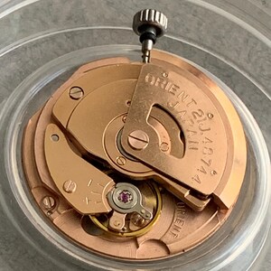 Puede incluir: Primer plano de un movimiento de reloj con engranajes y componentes dorados. El texto "ORIENT 21J JAPAN 48744" es visible. Una peque&ntilde;a corona plateada est&aacute; en la parte superior.