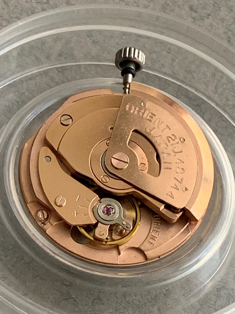 Puede incluir: Primer plano de un movimiento de reloj con engranajes y componentes dorados. El texto "ORIENT 21J JAPAN 48744" es visible. Una peque&ntilde;a corona plateada est&aacute; en la parte superior.