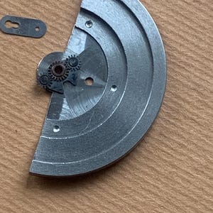 Pu&ograve; includere: Primo piano di componenti di orologio. Un componente metallico argentato a forma di semicerchio con tre piccoli fori e un meccanismo a ingranaggi. &Egrave; presente anche un piccolo pezzo di metallo rettangolare con un foro. I pezzi sono su una superficie marrone chiaro.