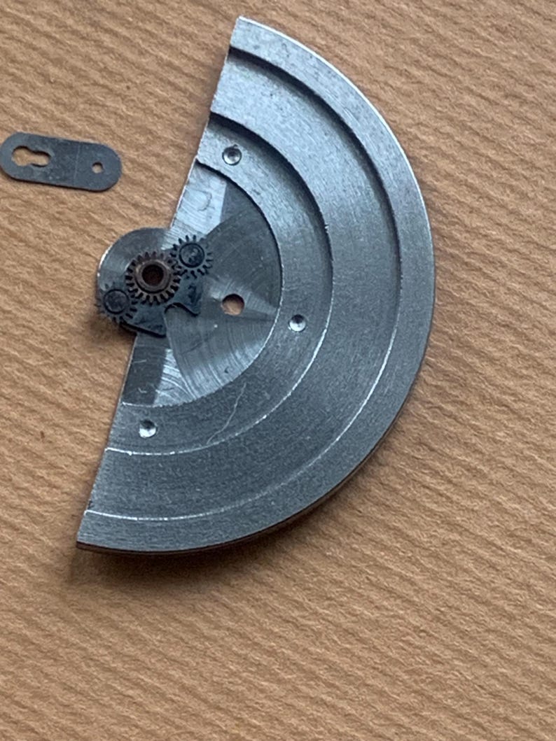 Pu&ograve; includere: Primo piano di componenti di orologio. Un componente metallico argentato a forma di semicerchio con tre piccoli fori e un meccanismo a ingranaggi. &Egrave; presente anche un piccolo pezzo di metallo rettangolare con un foro. I pezzi sono su una superficie marrone chiaro.