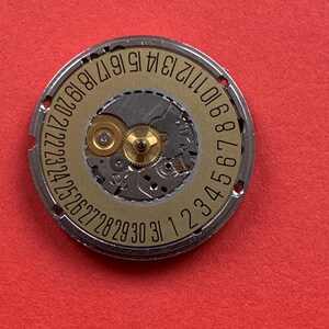 May include: Close-up of a watch movement with a gold-colored dial and silver-colored outer ring. The dial features the numbers 1 through 31, indicating the days of the month. The movement's intricate gears and components are visible.