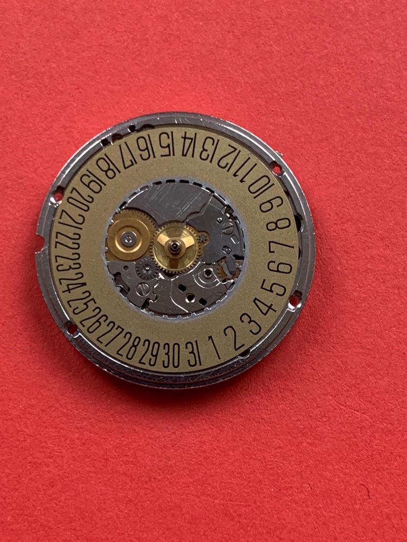 May include: Close-up of a watch movement with a gold-colored dial and silver-colored outer ring. The dial features the numbers 1 through 31, indicating the days of the month. The movement's intricate gears and components are visible.