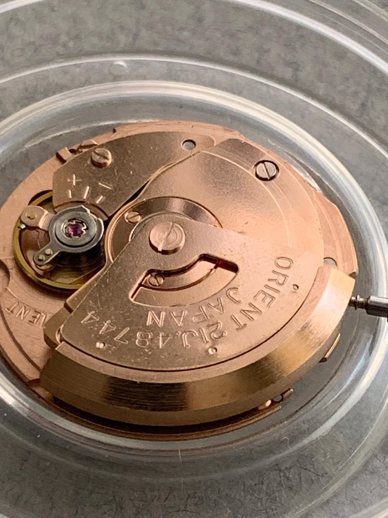 Puede incluir: Primer plano de un movimiento de reloj con engranajes y componentes dorados. El texto "ORIENT JAPAN 21J 48744" es visible en la placa principal. Una peque&ntilde;a joya roja est&aacute; engastada en el mecanismo.