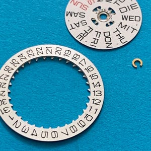 May include: Close-up of watch parts on a blue surface. The image shows a white day-of-the-week dial with black text and a red "SUN". A white date dial with numbers 1-31 and a small gold-colored clasp are also visible.