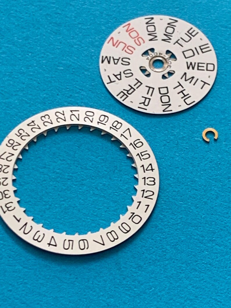 May include: Close-up of watch parts on a blue surface. The image shows a white day-of-the-week dial with black text and a red "SUN". A white date dial with numbers 1-31 and a small gold-colored clasp are also visible.