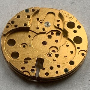 May include: Close-up of a gold-toned watch movement with intricate gears, holes, and mechanical components. The number "2571" is visible. The circular design suggests a vintage or antique timepiece.