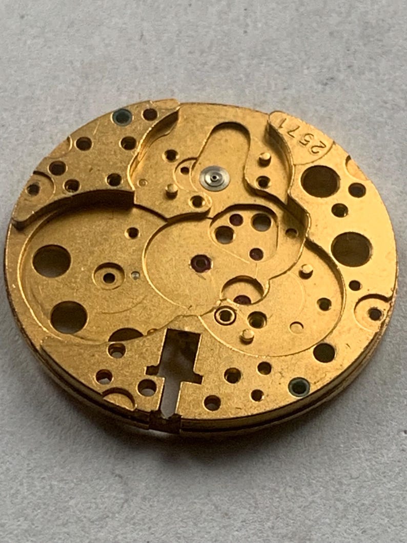 May include: Close-up of a gold-toned watch movement with intricate gears, holes, and mechanical components. The number "2571" is visible. The circular design suggests a vintage or antique timepiece.