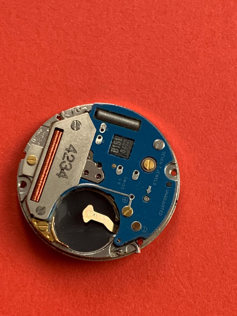 May include: A close-up of a watch movement with a blue circuit board and silver metal components. The number "4234" is printed on a silver component. The text "SEVEN 7 JEWELS" and "UNADJUSTED" are visible.