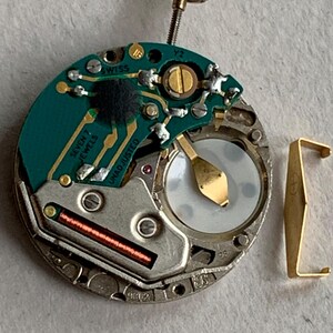 Puede incluir: Primer plano de un movimiento de reloj con una placa de circuito verde, componentes dorados y una peque&ntilde;a corona dorada. El movimiento tiene el texto "SWISS" y "SEVEN 7 JEWELS" impreso. Tambi&eacute;n se ve una pieza met&aacute;lica dorada separada.