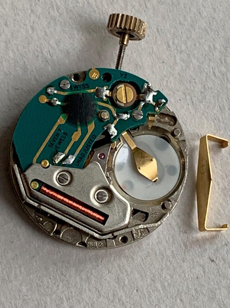Puede incluir: Primer plano de un movimiento de reloj con una placa de circuito verde, componentes dorados y una peque&ntilde;a corona dorada. El movimiento tiene el texto "SWISS" y "SEVEN 7 JEWELS" impreso. Tambi&eacute;n se ve una pieza met&aacute;lica dorada separada.