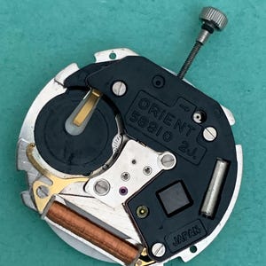 May include: A detailed view of a watch movement, featuring the text "ORIENT 58910 2J JAPAN". The mechanism has a black plate, silver parts, and a small silver crown. The background is a teal colour.
