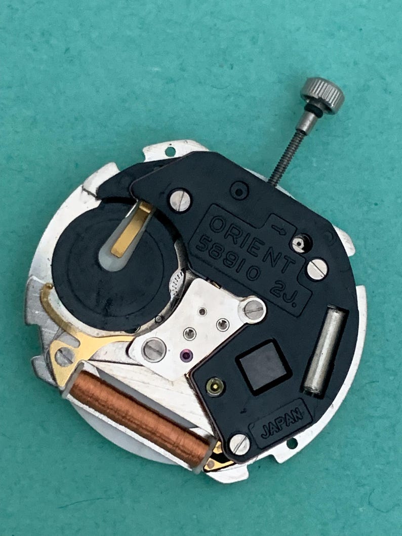 May include: A detailed view of a watch movement, featuring the text "ORIENT 58910 2J JAPAN". The mechanism has a black plate, silver parts, and a small silver crown. The background is a teal colour.