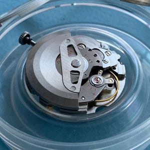 May include: A detailed view of a watch movement, showing its internal components. The silver-toned mechanism is partially open, displaying gears, screws, and a balance wheel. The watch parts are in a clear plastic container.