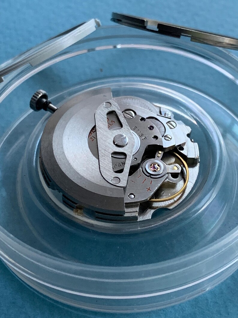 May include: A detailed view of a watch movement, showing its internal components. The silver-toned mechanism is partially open, displaying gears, screws, and a balance wheel. The watch parts are in a clear plastic container.
