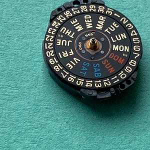 May include: A black watch calendar dial with gold-colored accents, displaying the days of the week in English and numbers 1-31. The dial has the text "555" and "0160" printed on it. The dial is set against a teal background.