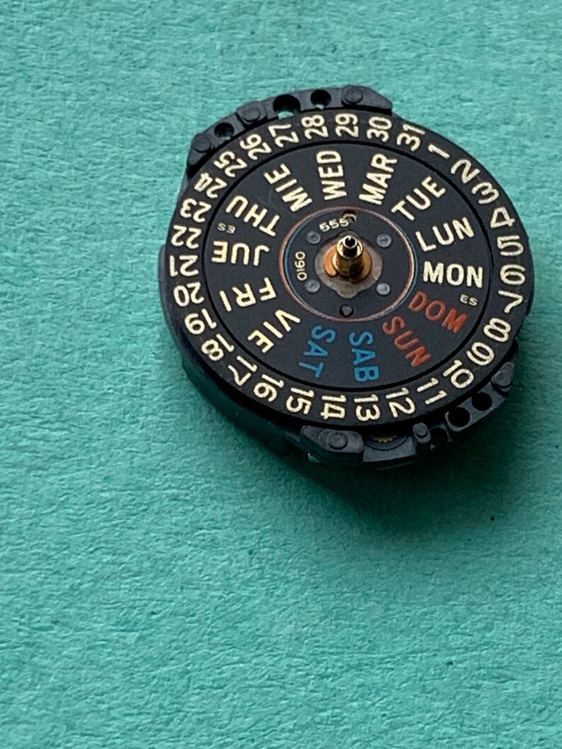 May include: A black watch calendar dial with gold-colored accents, displaying the days of the week in English and numbers 1-31. The dial has the text "555" and "0160" printed on it. The dial is set against a teal background.