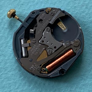 K&ouml;nnte beinhalten: Nahaufnahme eines Uhrwerks mit der Aufschrift "Jules Jurgensen 3W00 Japan". Das Uhrwerk hat eine goldfarbene Krone, eine kupferfarbene Spule und verschiedene kleine Metallkomponenten. Die W&ouml;rter "PUSH" und "UNADJUSTED" sind ebenfalls sichtbar.