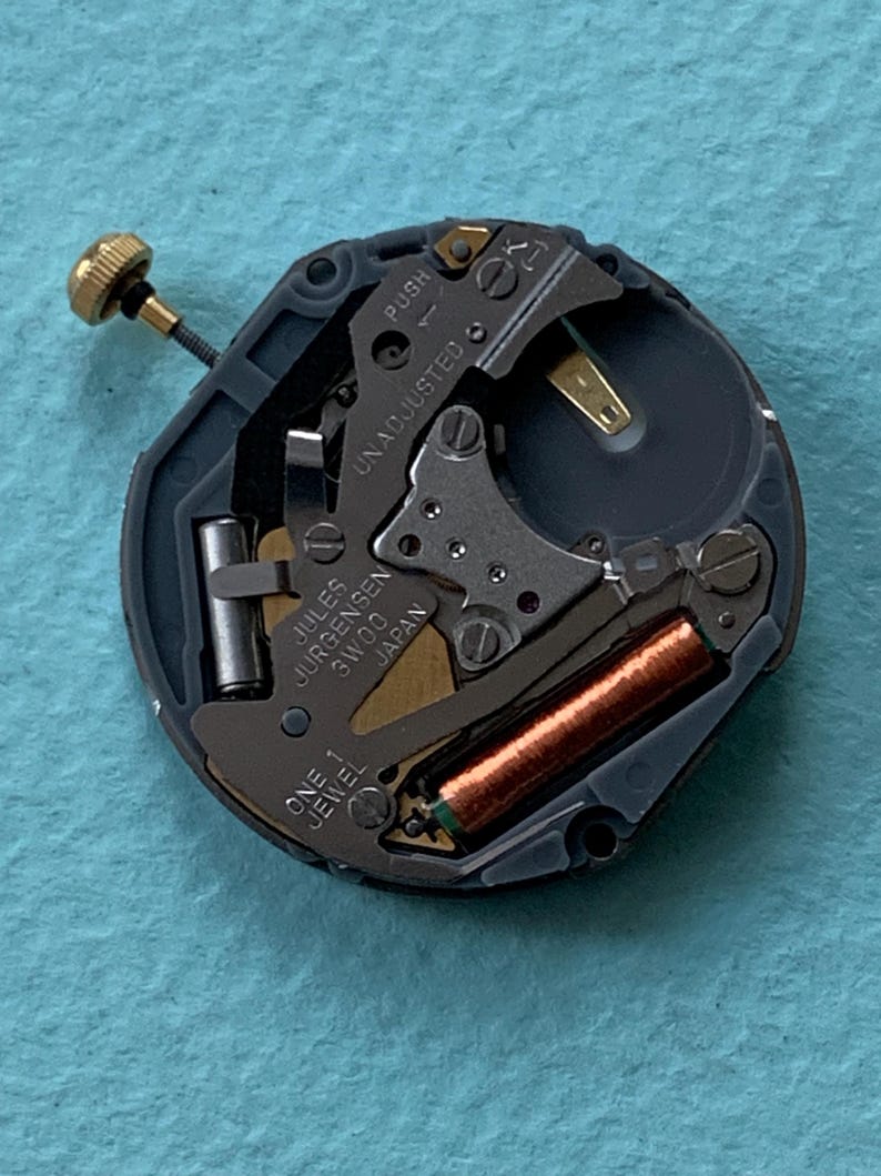 K&ouml;nnte beinhalten: Nahaufnahme eines Uhrwerks mit der Aufschrift "Jules Jurgensen 3W00 Japan". Das Uhrwerk hat eine goldfarbene Krone, eine kupferfarbene Spule und verschiedene kleine Metallkomponenten. Die W&ouml;rter "PUSH" und "UNADJUSTED" sind ebenfalls sichtbar.