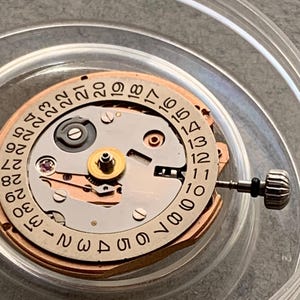Puede incluir: Primer plano de un movimiento de reloj con una rueda de fecha. El mecanismo presenta un dise&ntilde;o circular con n&uacute;meros del 1 al 31. El movimiento del reloj es de metal y tiene una corona.