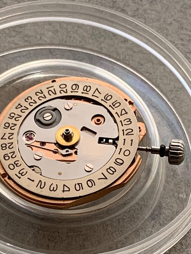Puede incluir: Primer plano de un movimiento de reloj con una rueda de fecha. El mecanismo presenta un dise&ntilde;o circular con n&uacute;meros del 1 al 31. El movimiento del reloj es de metal y tiene una corona.