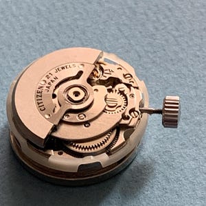 May include: A close-up of a Citizen watch movement with the text "CITIZEN 21 JEWELS JAPAN" visible. The watch mechanism is silver-toned with intricate gears and a crown. The background is a light blue surface.