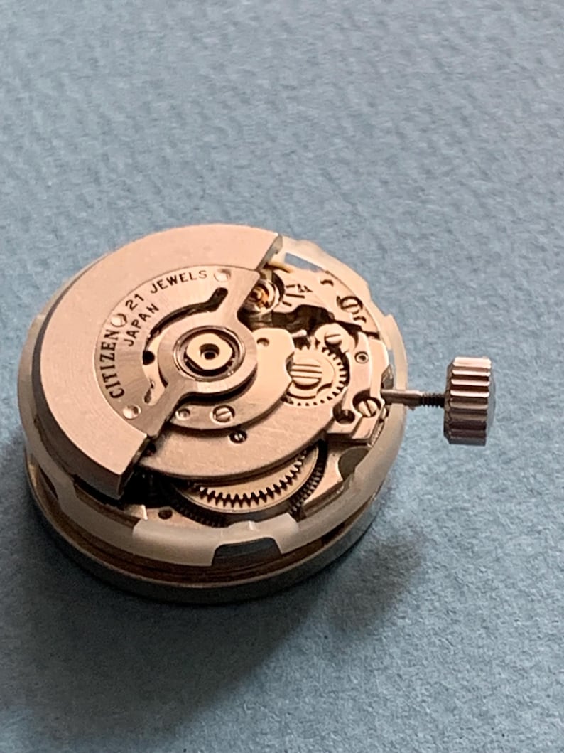 May include: A close-up of a Citizen watch movement with the text "CITIZEN 21 JEWELS JAPAN" visible. The watch mechanism is silver-toned with intricate gears and a crown. The background is a light blue surface.