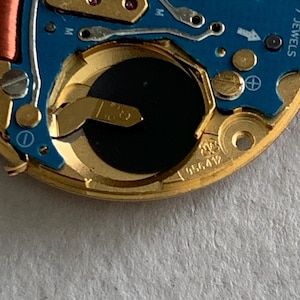 May include: Close-up of a watch movement with a blue circuit board and gold-colored metal components. The watch mechanism includes a battery, coils, and the text "SWISS V8 M". The serial number "956412" is visible.
