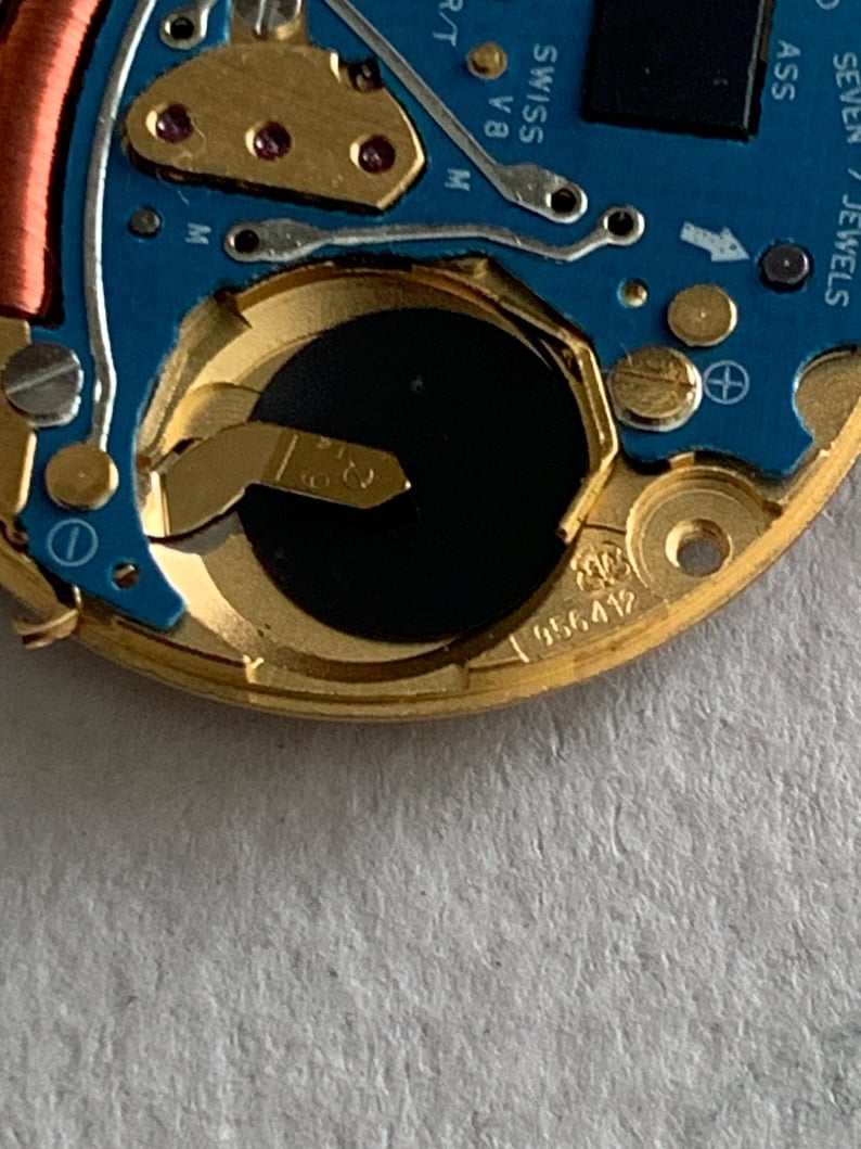 May include: Close-up of a watch movement with a blue circuit board and gold-colored metal components. The watch mechanism includes a battery, coils, and the text "SWISS V8 M". The serial number "956412" is visible.