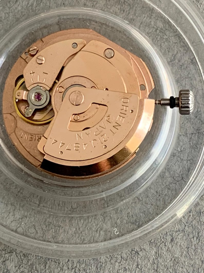 Puede incluir: Primer plano de un movimiento de reloj con engranajes y componentes dorados. Las palabras "ORIENT JAPAN" son visibles en una pieza curva. Una peque&ntilde;a joya roja est&aacute; engastada en el centro. El movimiento est&aacute; dentro de un recipiente de pl&aacute;stico transparente.