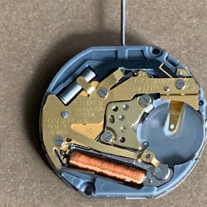 May include: Close-up of a Citizen watch movement. The watch mechanism features gold-toned metal components, a copper-colored coil, and a gray plastic casing. The text "CITIZEN WATCH CO." is visible.