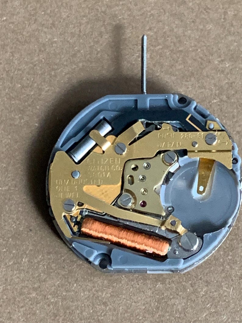 May include: Close-up of a Citizen watch movement. The watch mechanism features gold-toned metal components, a copper-colored coil, and a gray plastic casing. The text "CITIZEN WATCH CO." is visible.