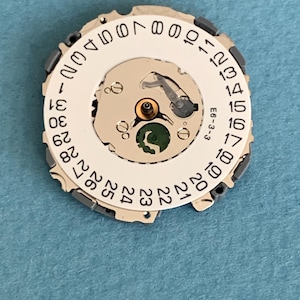 May include: A watch movement component with a white circular dial featuring black numbers from 1 to 31. The dial has a central mechanism with a green element and metallic components. The text "E6-3-3" is visible.