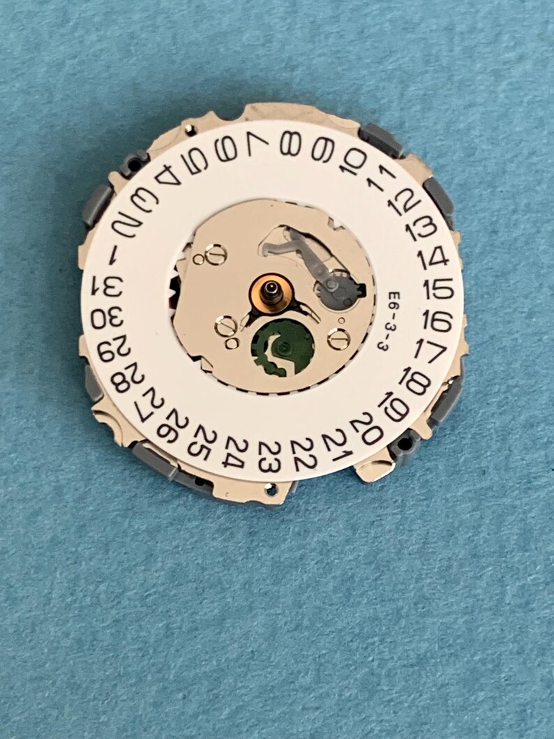 May include: A watch movement component with a white circular dial featuring black numbers from 1 to 31. The dial has a central mechanism with a green element and metallic components. The text "E6-3-3" is visible.