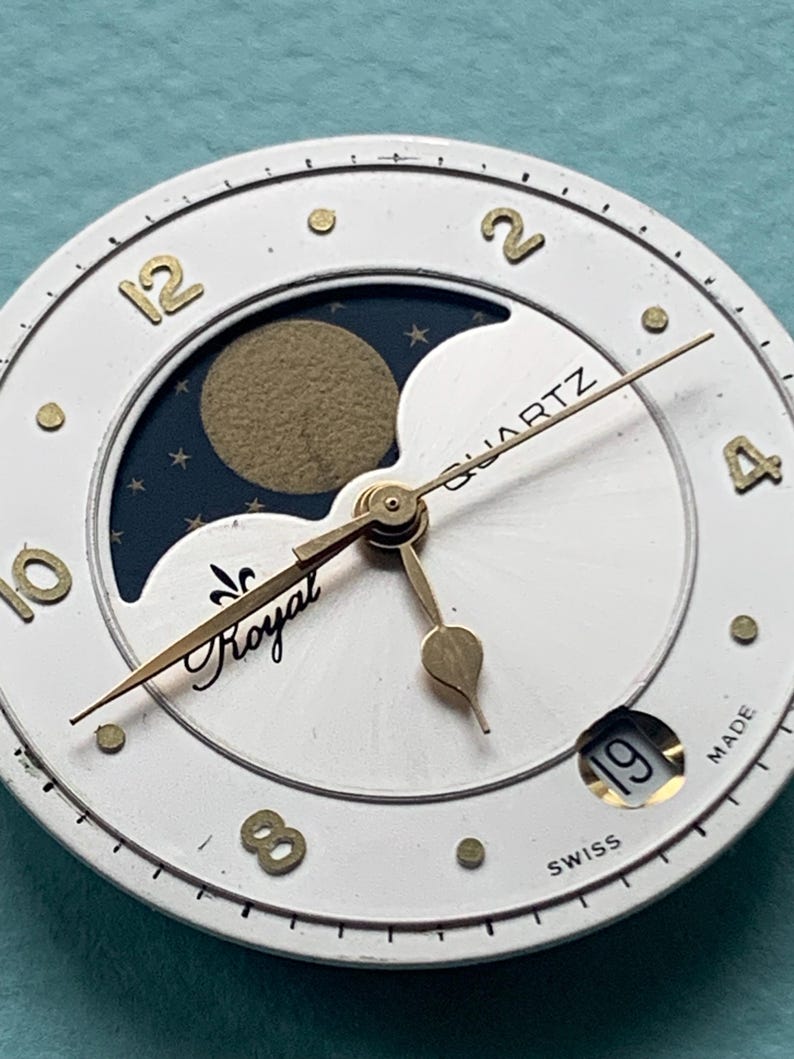 Op de afbeelding: Close-up van een witte wijzerplaat met goudkleurige wijzers en cijfers. De wijzerplaat heeft een maanfase-indicator met een gouden maan en sterren. Het woord "Royal" is in cursief geschreven.