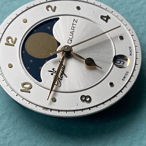 Op de afbeelding: Een horlogekast met een witte buitenring en een zilveren midden. Het heeft gouden wijzers, cijfers en een maanfase-indicator. De teksten "QUARTZ" en "Royal" zijn zichtbaar. Het datumvenster toont het nummer 19.