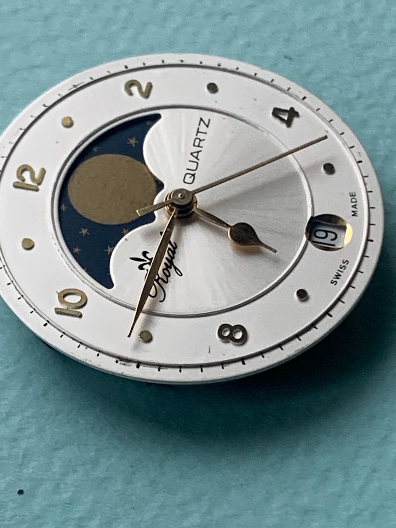 Op de afbeelding: Een horlogekast met een witte buitenring en een zilveren midden. Het heeft gouden wijzers, cijfers en een maanfase-indicator. De teksten "QUARTZ" en "Royal" zijn zichtbaar. Het datumvenster toont het nummer 19.