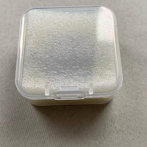 May include: A small, clear plastic box with a hinged lid. The interior is filled with a light-colored, textured foam. The box is square with rounded corners and a secure latch.